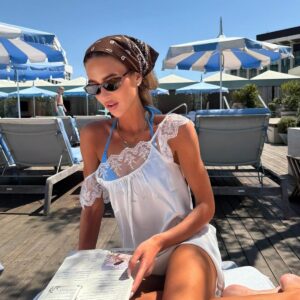 Девушка в узких черных солнцезащитных очках, коричневом платке и белом летнем платье у бассейна