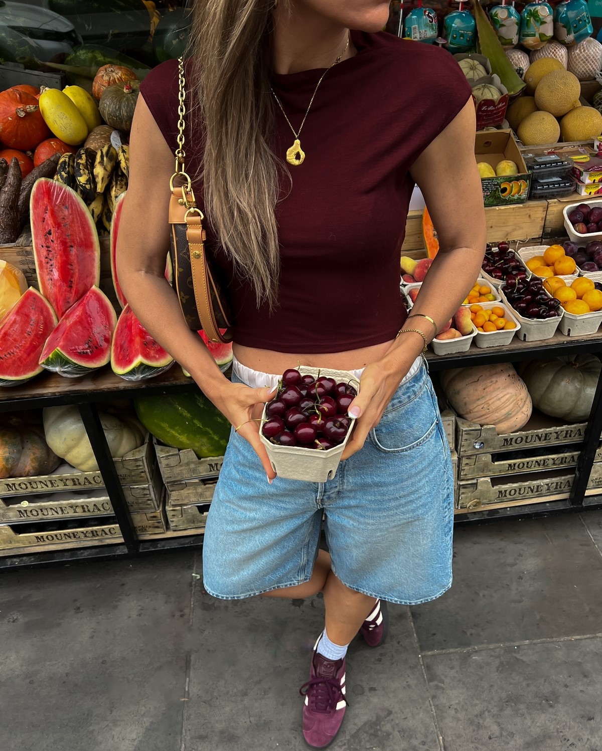Женщина в бордовом топе и джинсовых шортах jorts выбирает черешню на рынке