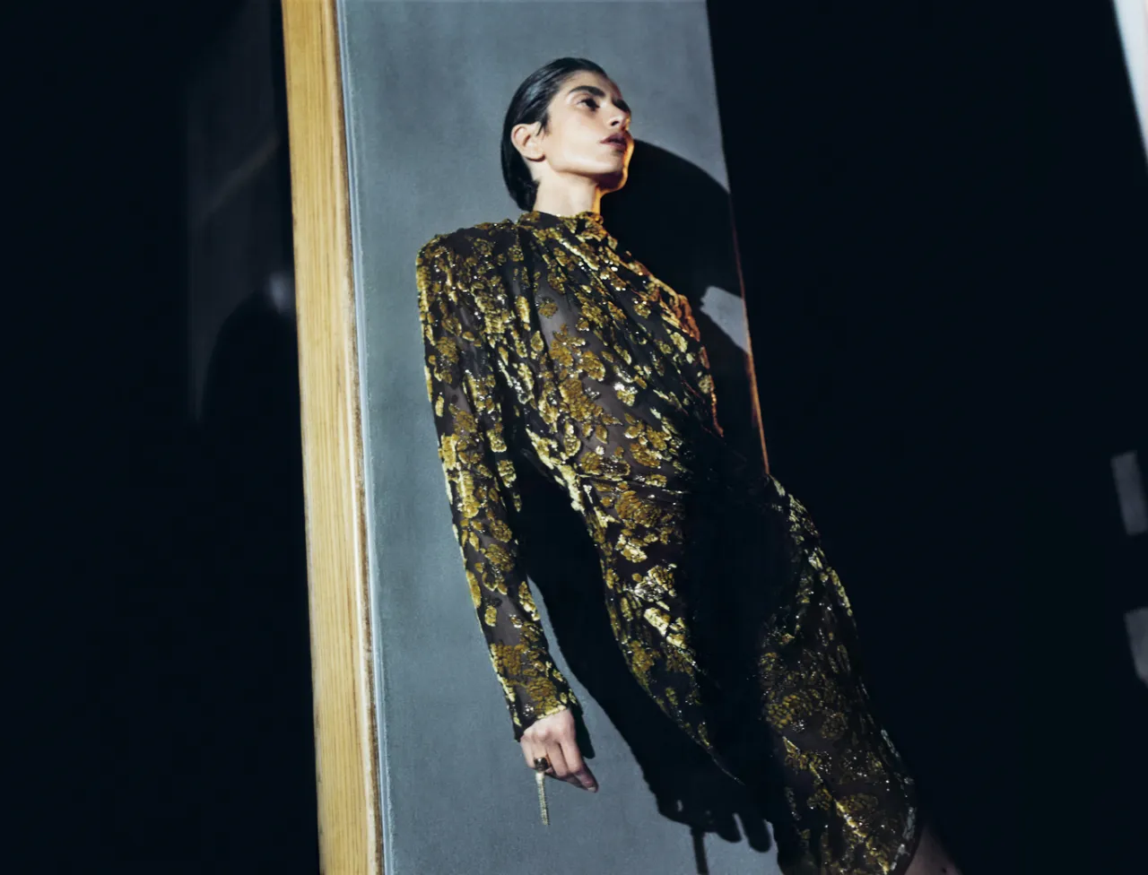 Кадр кампании Balmain: модель в черном платье с золотистым цветочным декором у зеркала
